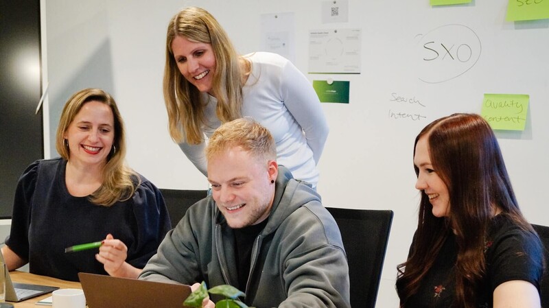 Vier Personen sitzen vor einem Laptop am Tisch im Konferenzraum