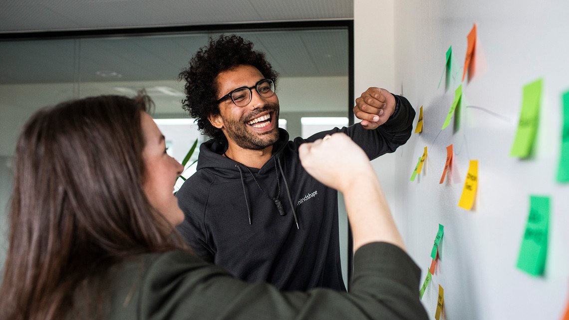 Jobs bei mindshape: Zwei Personen kleben Post-its an die Wand.