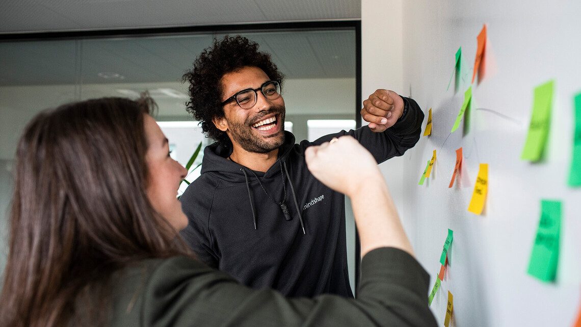 Jobs bei mindshape: Unsere Benefits verraten, warum sich eine Bewerbung lohnt. Jobs bei mindshape: Zwei Personen kleben Post-its an die Wand.