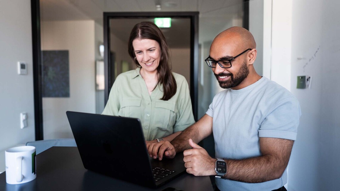 Case Studies: zwei Kolleg:innen stehen vor einem Tisch im Meeting-Raum und schauen auf Laptop