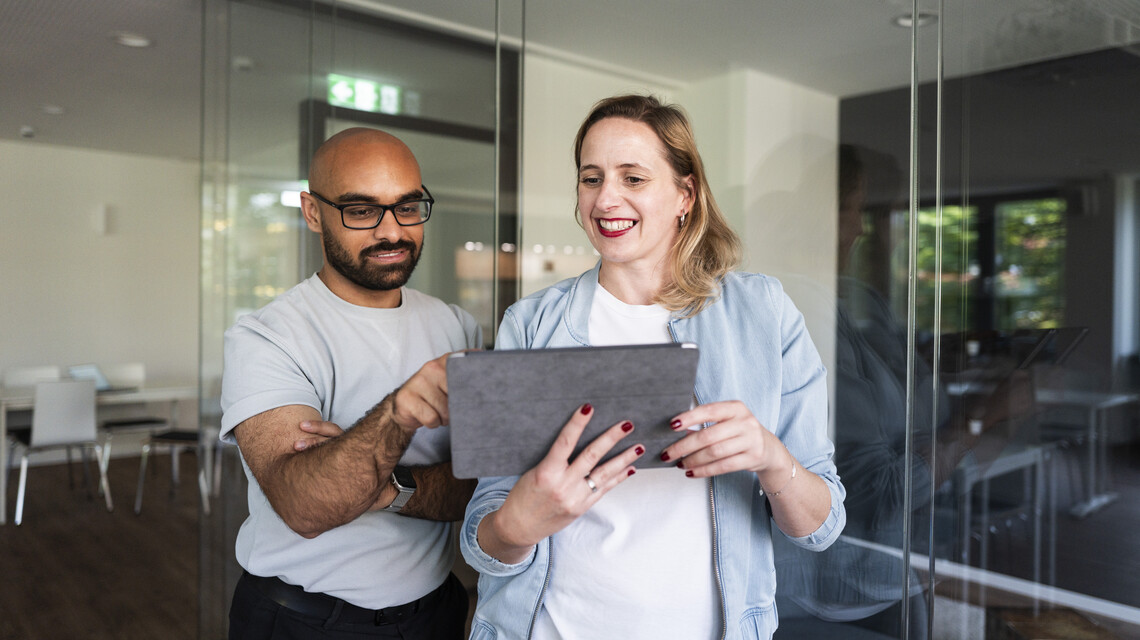 Online Marketing Agentur: zwei Kolleg:innen schauen vor einem verglasten Meeting-Raum auf ein Tablet